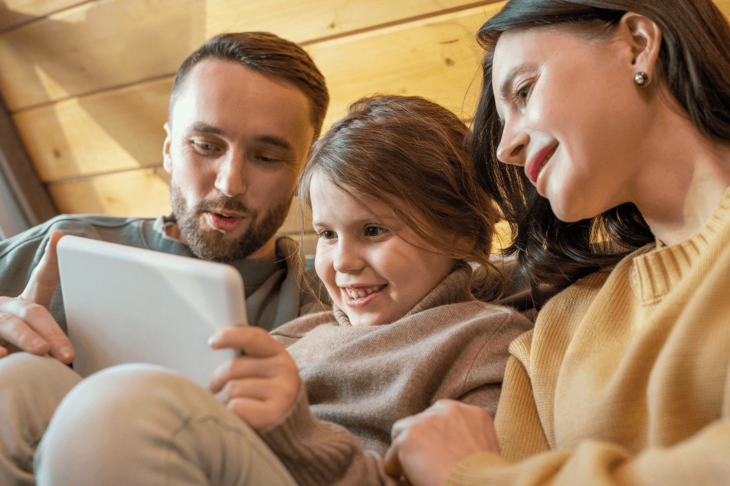 Eine junge Familie – Vater, Mutter und Tochter – sitzt eng zusammen vor einer hellen Holzwand und schaut lächelnd auf ein gemeinsames Tablet.